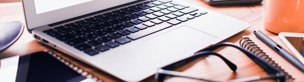 Closeup of a laptop keyboard with other devices nearby as well as classes, notebooks, and pens.