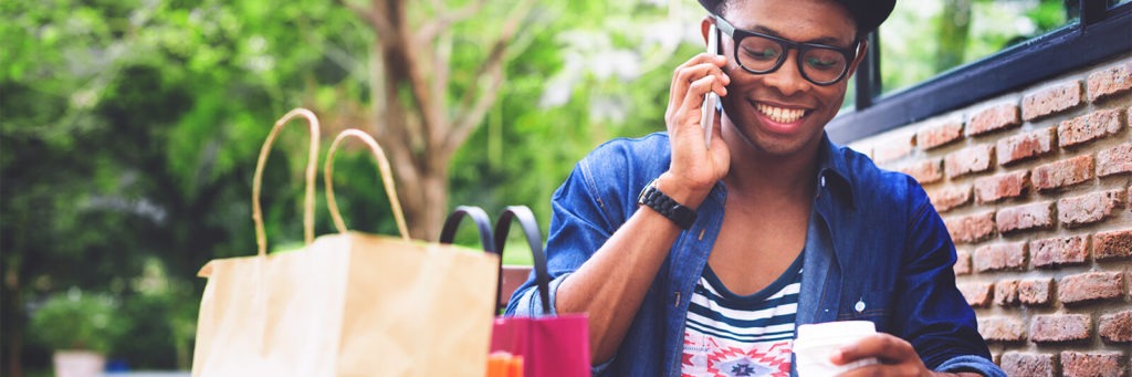 Young person sitting down with a coffee next to a couple of shopping bags, smiling and listening to a phone.