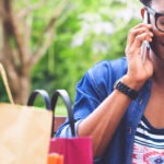 Young person sitting down with a coffee next to a couple of shopping bags, smiling and listening to a phone.