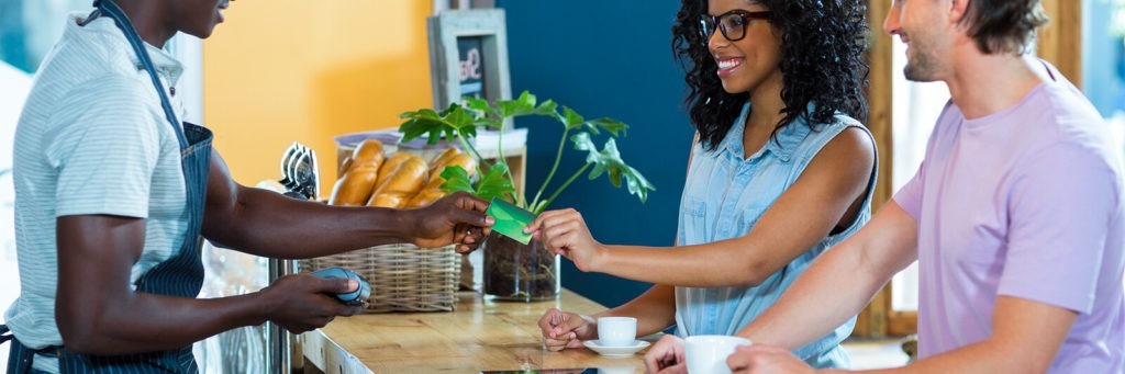 Woman with black curly hair and black frame glasses passes a green credit card over to a barista while smile in exchange for a coffee.