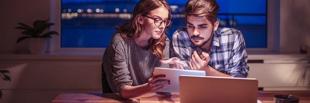 A young man and woman sit at a table and are bathed in a laptop light in thier living room while they look at a paper together.