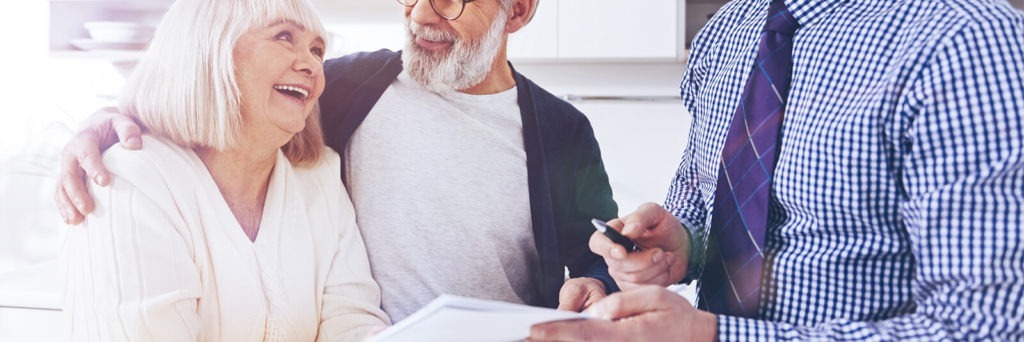 Older man and woman with white hair smile at one another next to a man in a gingham dress shit holding a pen and paper.