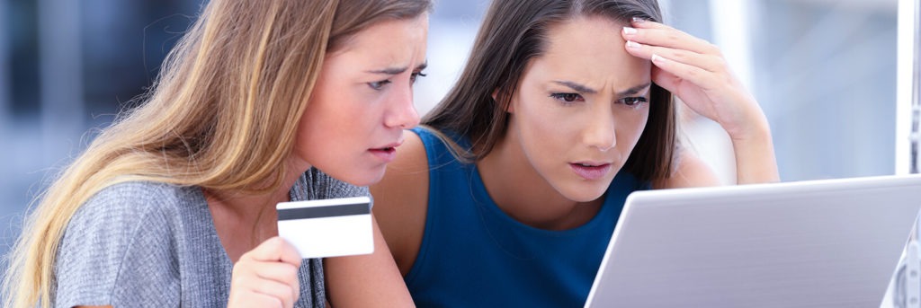 Two young women look worriedly at a laptop screen while one of them holds a credit card in the air.