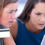 Two young women look worriedly at a laptop screen while one of them holds a credit card in the air.